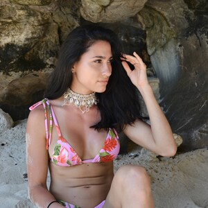 Puede incluir: Una mujer con un top de bikini floral rosa y una parte de abajo a juego. Est&aacute; sentada en una playa de arena con un acantilado rocoso detr&aacute;s de ella. Lleva un collar de conchas.