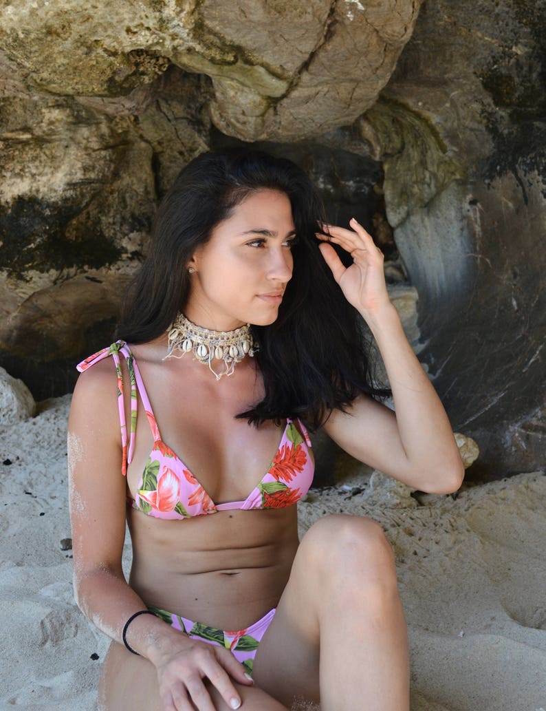 Puede incluir: Una mujer con un top de bikini floral rosa y una parte de abajo a juego. Est&aacute; sentada en una playa de arena con un acantilado rocoso detr&aacute;s de ella. Lleva un collar de conchas.