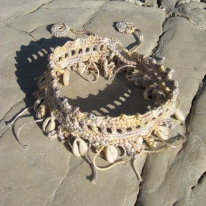Puede incluir: Una diadema de crochet beige y marr&oacute;n con conchas blancas unidas. La diadema est&aacute; sobre una superficie rocosa gris claro.