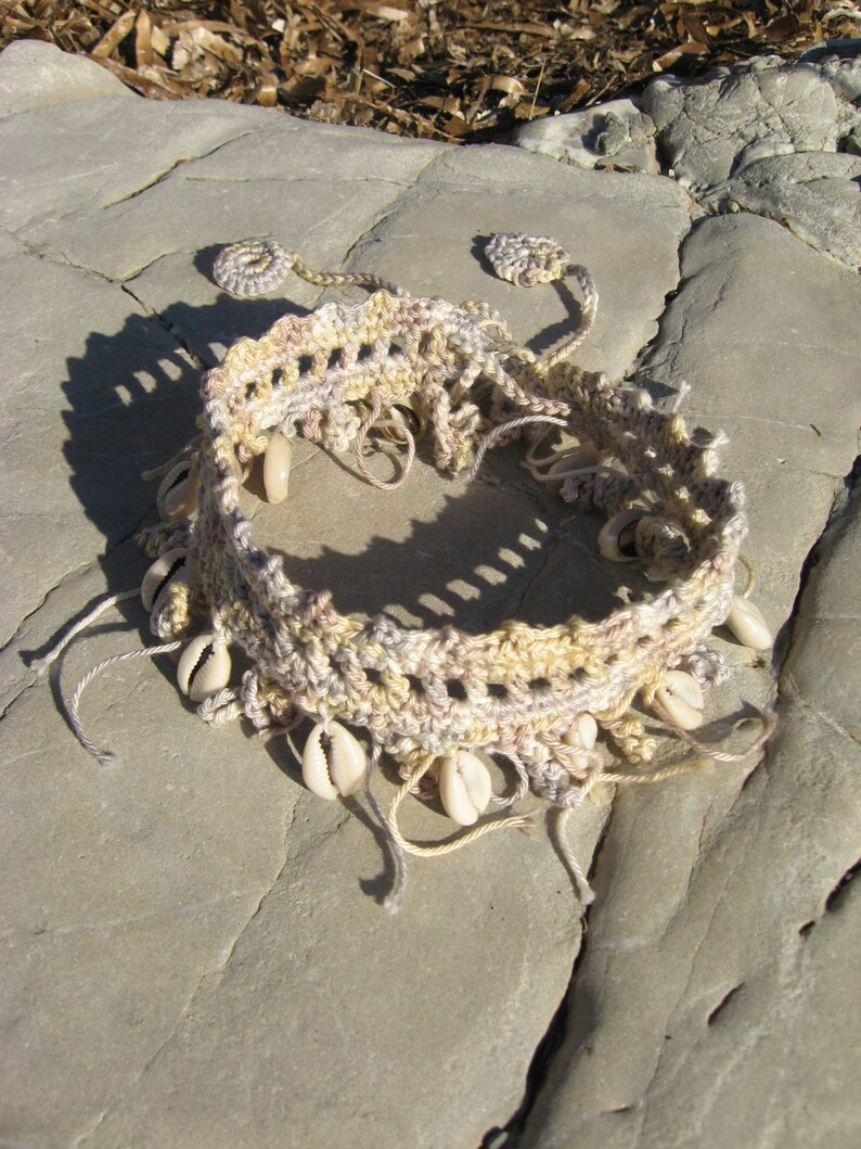 Puede incluir: Una diadema de crochet beige y marr&oacute;n con conchas blancas unidas. La diadema est&aacute; sobre una superficie rocosa gris claro.