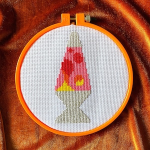 May include: A cross-stitch embroidery hoop featuring a colourful lava lamp design. The lamp is pink, orange and yellow with a grey base.