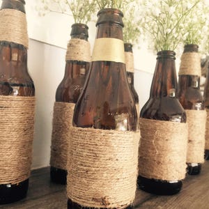 May include: A row of brown glass bottles wrapped in twine with white flowers in the top. The bottles are arranged on a wooden surface.