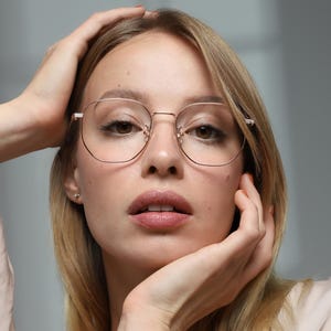 Op de afbeelding: Een persoon met een ronde bril met een gouden montuur. De bril heeft een dun montuur en een moderne, geometrische vorm. De persoon heeft blond haar en draagt een lichtgekleurde blouse. Ze houdt haar handen naar haar hoofd.