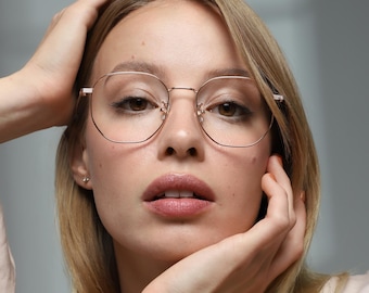 May include: A woman wearing round, gold-rimmed eyeglasses. The glasses have a thin frame and a modern, geometric shape. The woman has blonde hair and is wearing a light-colored blouse. She is holding her hands up to her head.