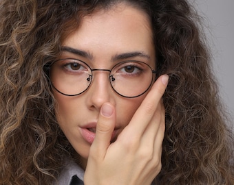 Könnte beinhalten: Eine Frau mit lockigem braunen Haar, die eine runde Brille mit schwarzem Rahmen und ein graues Hemd mit Knöpfen trägt.