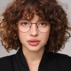 May include: A woman with curly brown hair wearing black round-framed glasses and a black v-neck top.