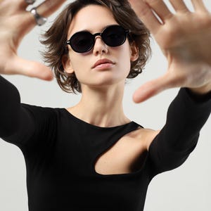 Woman wearing round black sunglasses with dark lenses and tortoiseshell temples, posing with her hands framing her face.