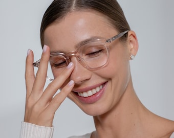 May include: A person wearing clear-framed eyeglasses with a square shape. The frames have silver accents on the temples. The person is smiling and wearing a white sweater.