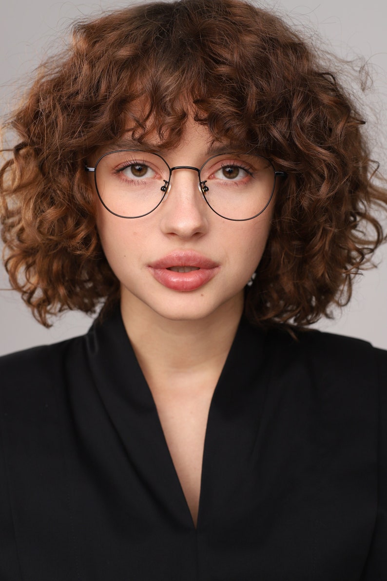 May include: A woman with curly brown hair wearing black round-framed glasses and a black v-neck top.