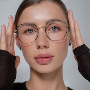 May include: Geometric eyeglasses with gold rims and black frames. The glasses have a double bridge. The lenses are clear. The style is modern and sophisticated. The glasses are being worn by a person.
