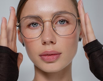 May include: Gold-rimmed, geometric eyeglasses with black frames. The glasses have a double bridge and are worn by a person. The lenses are clear. The overall style is modern and sophisticated.