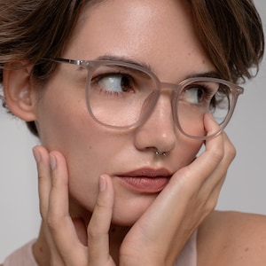 May include: A close-up of eyeglasses with a light pink frame. The square-shaped glasses have a thin silver metal bridge. The person is wearing a light pink tank top and has a nose ring. The person is touching their face with their hands.