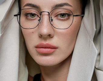 May include: A close-up portrait features a person wearing black, round-framed eyeglasses. The person is wearing a black top and a light beige jacket. The background is a soft, neutral color.