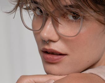 May include: A close-up shot of a person wearing clear-framed eyeglasses. The glasses have a square shape. The person has short, brown hair and a nose ring. The person is resting their chin on their hand.