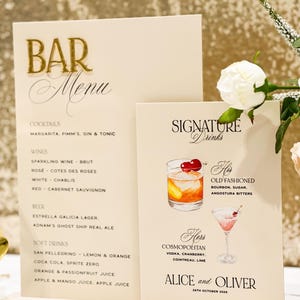 May include: Two cream-coloured wedding bar menus. The larger menu lists cocktails, wines, beer, and soft drinks. The smaller menu features signature drinks, including an Old Fashioned and a Cosmopolitan, with the names "Alice and Oliver" and the date "26th October 2026."