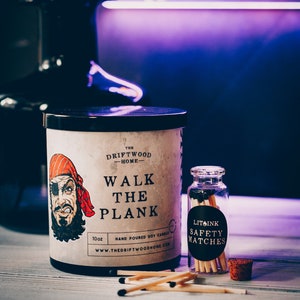 May include: A 10 ounce soy candle in a black and white jar with a pirate illustration and the text "The Driftwood Home - Walk the Plank". The candle is next to a small glass jar of matches with the text "Litsink Safety Matches".