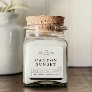 May include: A clear glass jar candle with a cork lid. The label reads "The Driftwood Home - Canyon Sunset 8oz Hand Poured Soy Candle www.thedriftwoodhome.com".