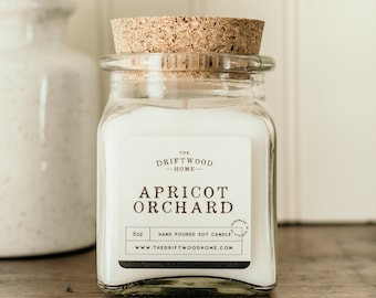 May include: A clear glass jar candle with a cork lid. The label reads "The Driftwood Home" and "Apricot Orchard". The candle is 8 oz and hand-poured soy candle. The website address is www.thedriftwoodhome.com.