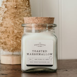 May include: A clear glass jar candle with a cork lid. The label reads "The Driftwood Home" and "Toasted Marshmallow". The candle is hand-poured soy and is 8 oz.