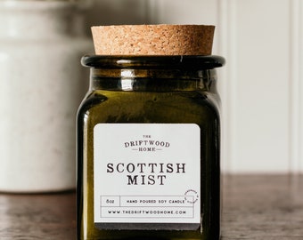 May include: A dark green glass jar with a cork lid and a white label that reads "The Driftwood Home - Scottish Mist". The label also includes the text "8oz Hand Poured Soy Candle" and "www.thedriftwoodhome.com".
