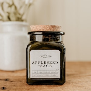 May include: A dark green glass jar with a cork lid and a white label. The label reads "The Driftwood Home" and "Appleseed + Sage". It also says "6oz Hand Poured Soy Candle" and "www.thedriftwoodhome.com".
