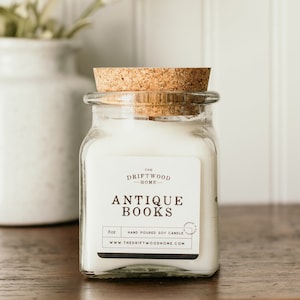 May include: A white soy candle in a clear glass jar with a cork lid. The label reads "The Driftwood Home" and "Antique Books". The candle is 8 oz and hand-poured.