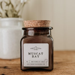 May include: A brown glass jar candle with a cork lid. The label reads "The Driftwood Home" and "Muscat Bay". The candle is hand-poured soy and is 6 oz.