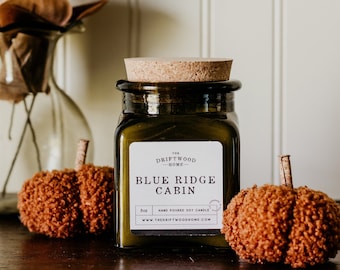 May include: A brown glass candle jar with a cork lid. The label reads "The Driftwood Home - Blue Ridge Cabin" and "8oz Hand Poured Soy Candle - www.thedriftwoodhome.com". The jar is sitting on a wooden surface with two brown fuzzy pumpkins.