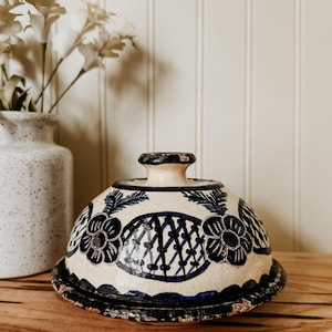 May include: A ceramic butter dish with a dome-shaped lid featuring a cream-colored base and hand-painted dark blue floral and lattice patterns. The lid has a central knob handle, and the base plate has a dark blue scalloped edge with a distressed finish.