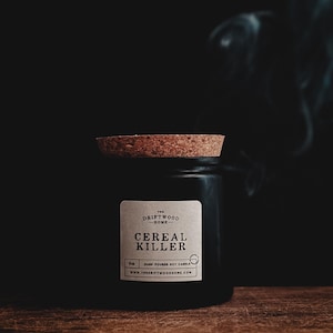 May include: A black jar candle with a cork lid, labeled "Cereal Killer." The label also includes "The Driftwood Home" and "Hand Poured Soy Candle." Smoke rises in the background, and the candle sits on a wooden surface.