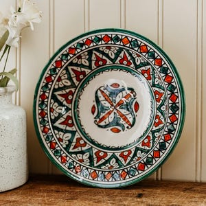 May include: A decorative ceramic plate with a detailed, colorful design. The plate features a white base with a central pattern and a border of green, red, and orange geometric shapes. The rim is a solid green. The plate is on a wooden surface.