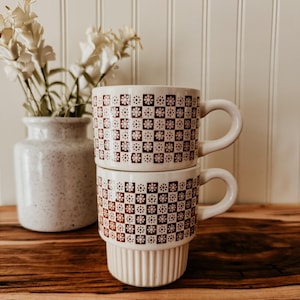 May include: Two stacked ceramic mugs featuring a repeating dark brown checkerboard pattern with small white floral icons in each square. The top mug has a smooth cylindrical body, while the bottom mug features a ribbed base design.