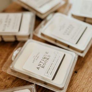 May include: A close-up of several wax melts in clear plastic containers. The wax melts are labeled with the text "The Driftwood Home" and "Antique Books" with the website address "www.thedriftwoodhome.com".