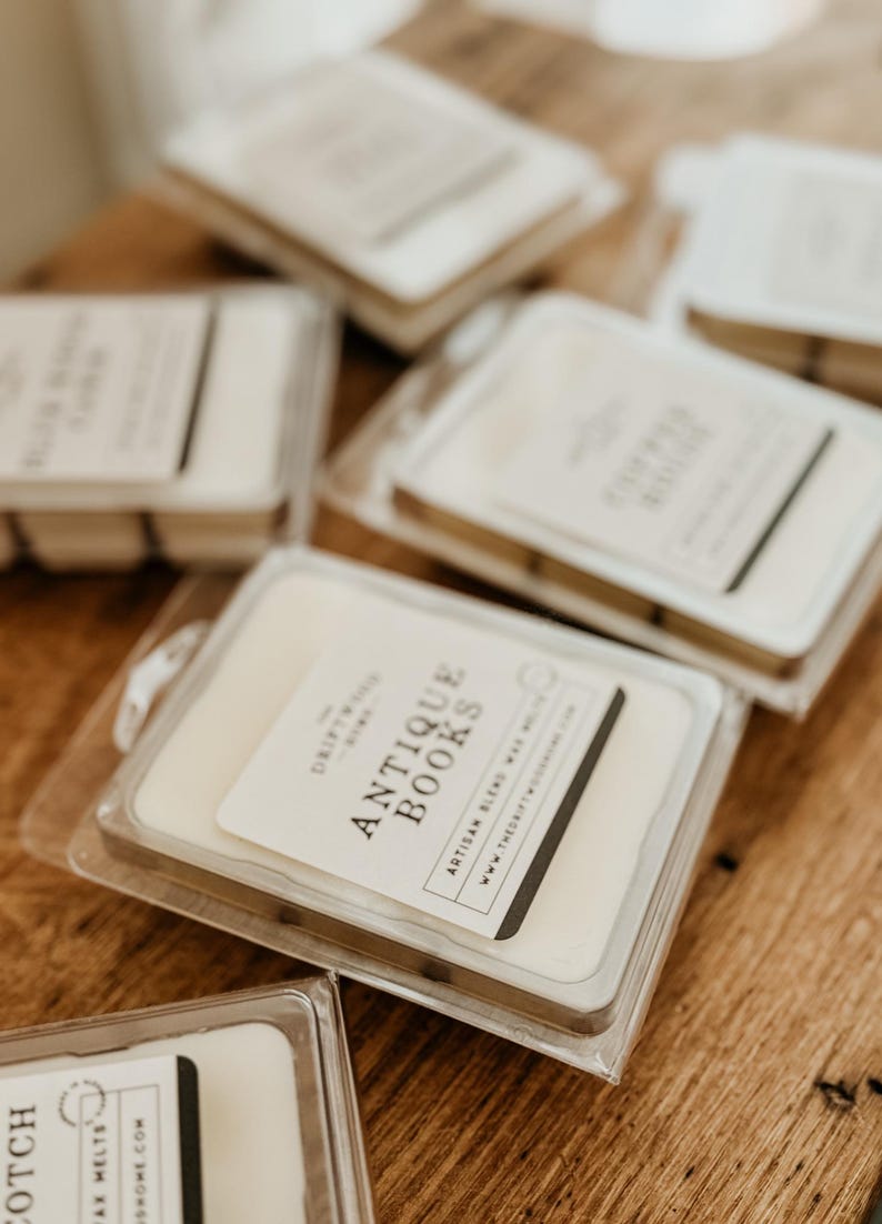 May include: A close-up of several wax melts in clear plastic containers. The wax melts are labeled with the text "The Driftwood Home" and "Antique Books" with the website address "www.thedriftwoodhome.com".