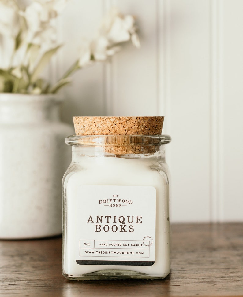 May include: A white soy candle in a clear glass jar with a cork lid. The label reads "The Driftwood Home" and "Antique Books". The candle is 8 oz and hand-poured.