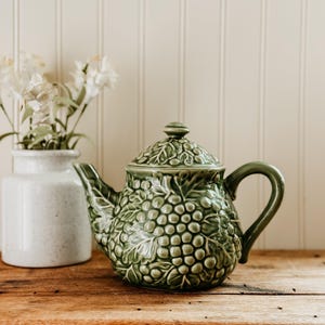 May include: A green ceramic teapot with a grape design, featuring a lid and curved handle. The teapot sits on a wooden surface next to a white vase with white flowers. The background is a white paneled wall.