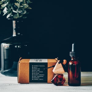 May include: A brown cardboard box with a list of scents, a wooden car air freshener with a glass bottle, and a glass bottle of essential oil. The scents listed on the box include Caribbean Teakwood, Vanilla, Summer Rain, Beach Vibes, Sea Salt Orchid, Driftwood, Coconut Pineapple, Sweet Lemongrass, Grainsacke, and Lavender Vanilla.