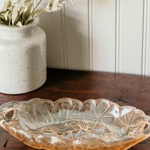 May include: A vintage, oval-shaped glass serving dish with two handles. The dish is made of clear glass with a frosted, iridescent finish. The dish has a floral design etched into the glass.