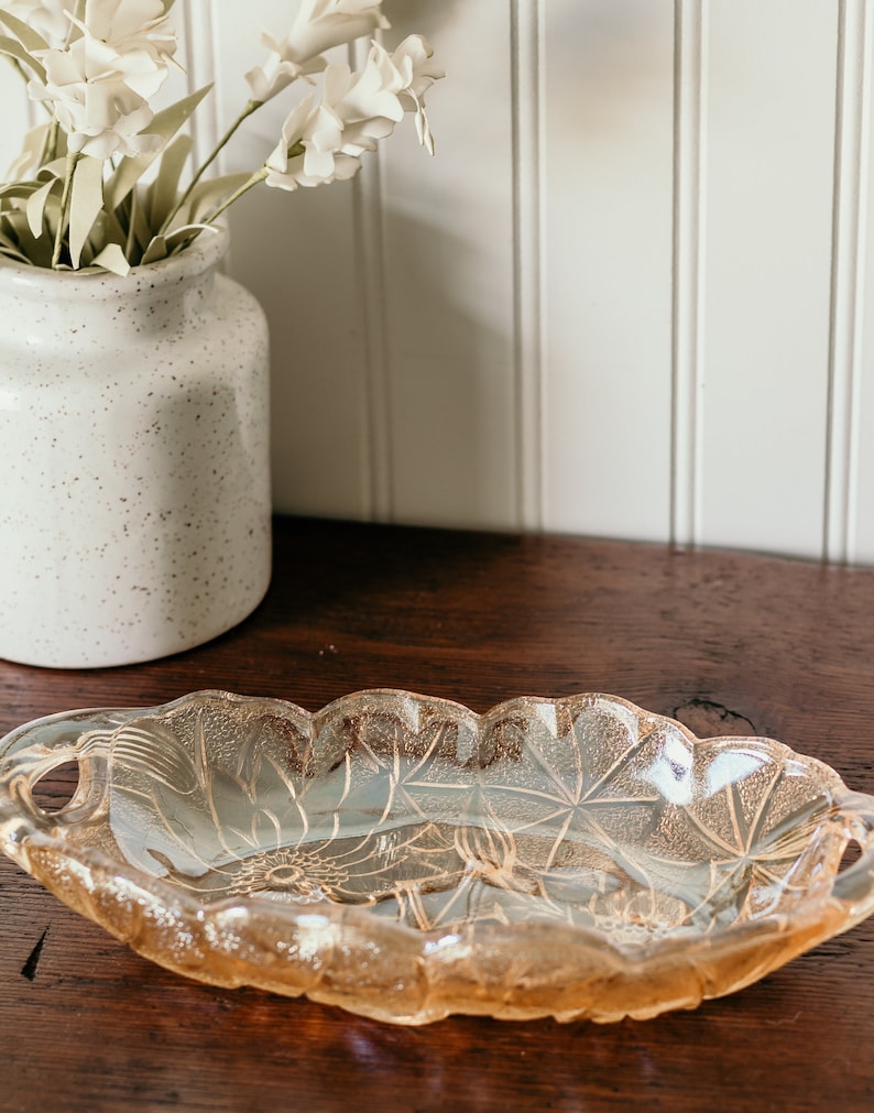 May include: A vintage, oval-shaped glass serving dish with two handles. The dish is made of clear glass with a frosted, iridescent finish. The dish has a floral design etched into the glass.