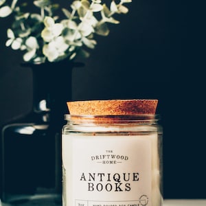 May include: A white candle jar with a cork lid. The label reads "The Driftwood Home - Antique Books" and "16oz Hand Poured Soy Candle". The website address is "www.thedriftwoodhome.com".