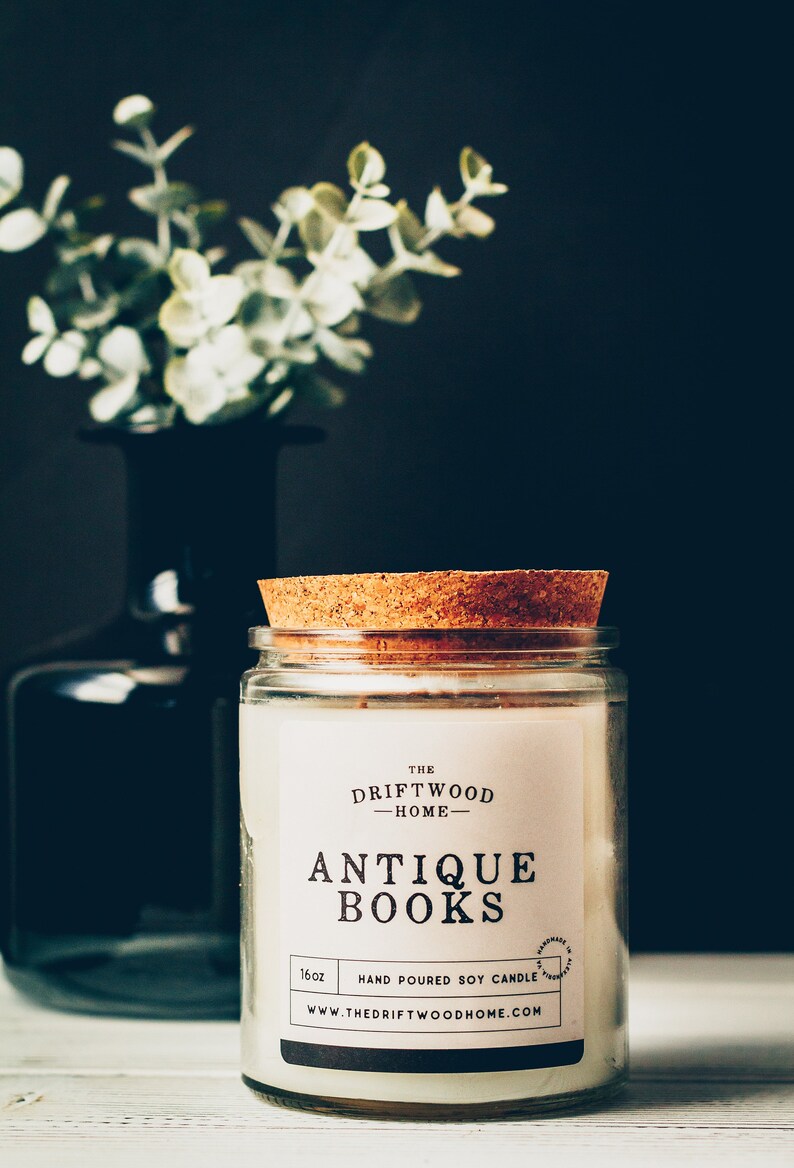 May include: A white candle jar with a cork lid. The label reads "The Driftwood Home - Antique Books" and "16oz Hand Poured Soy Candle". The website address is "www.thedriftwoodhome.com".