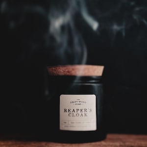 May include: A black glass jar candle with a cork lid, labeled "Reaper's Cloak" from The Driftwood Home. Wisps of smoke rise from the candle against a dark background. The candle sits on a wooden surface.
