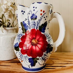 May include: A white ceramic pitcher featuring a hand-painted floral design with a large central red flower, blue and green leaves, and small orange accents. The pitcher has a curved handle, a scalloped rim, and a glossy finish on the exterior surface.
