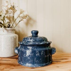 May include: A round, dark blue ceramic lidded casserole dish featuring a white marbled or bubble-like pattern across its surface. The vessel includes two side handles and a small knob on the lid, showcasing a glossy finish and a structured design.