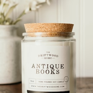May include: A clear glass jar candle with a cork lid. The label reads "The Driftwood Home - Antique Books" and "Hand Poured Soy Candle". The candle is 16 oz and is handmade in the USA.