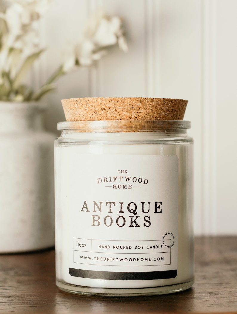 May include: A clear glass jar candle with a cork lid. The label reads "The Driftwood Home - Antique Books" and "Hand Poured Soy Candle". The candle is 16 oz and is handmade in the USA.
