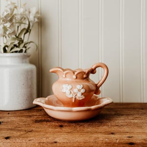 May include: A peach-colored ceramic pitcher and basin set with white floral accents. The pitcher has a curved handle and a scalloped rim. The basin has a matching scalloped edge. The set is displayed on a wooden surface.