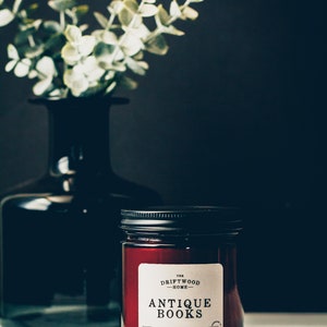 May include: A brown glass candle jar with a black lid and a white label that reads "The Driftwood Home - Antique Books - Hand Poured Soy Candle - 8oz - www.thedriftwoodhome.com".