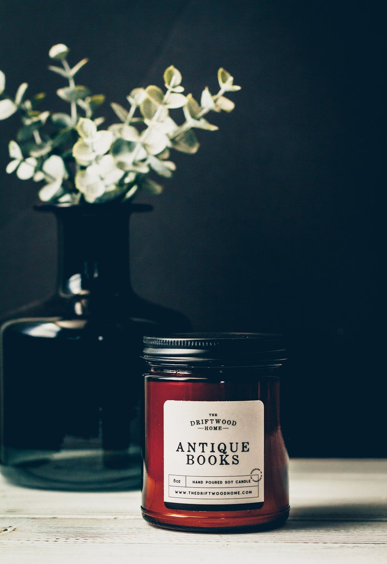 May include: A brown glass candle jar with a black lid and a white label that reads "The Driftwood Home - Antique Books - Hand Poured Soy Candle - 8oz - www.thedriftwoodhome.com".