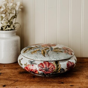 May include: A round, lidded trinket box with a floral design. The box features a light blue background with red and yellow flowers. The box is sitting on a wooden surface.
